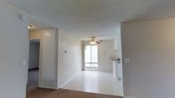This single-floor residence offers a clean and functional layout designed for comfortable everyday living. The main living area is open and airy, bathed in natural light streaming through a large window that anchors the far wall and provides views to the outside. A ceiling fan with an integrated light fixture is centrally positioned overhead, contributing to both comfort and airflow throughout the space. The floors transition from carpet to a smooth hard-surface finish near the kitchen and window area, adding subtle visual variety to the open floor plan. The walls are finished in a neutral white palette, complemented by a textured ceiling that runs throughout. A floor vent is integrated into the baseboard area, indicating a built-in climate control system. The kitchen is accessible directly from the living area, partially visible with white cabinetry and a countertop, suggesting a practical and connected layout ideal for daily use. The overall ambiance of the space is bright, understated, and move-in ready, offering a versatile canvas that can be personalized to a variety of tastes and styles. The open sightlines between the living and kitchen areas enhance the sense of space and ease of movement throughout the home.