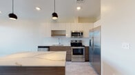 This contemporary single-floor residence showcases a thoughtfully designed open layout that emphasizes clean lines and modern finishes throughout. The kitchen serves as the centerpiece of the space, featuring a striking two-tone cabinetry scheme that pairs flat-panel white upper cabinets with warm wood-grain lower cabinets. A standout quartz-topped island with natural veining provides generous prep and gathering space, illuminated by sleek matte black pendant lights overhead. The kitchen is fully equipped with a stainless steel appliance suite, including a French door refrigerator, electric range with over-the-range microwave, and dishwasher, all set against a classic white tile backsplash. Light-toned flooring runs through the main living and kitchen areas, enhancing the sense of openness. The bathroom offers a clean, minimalist aesthetic with a glass-enclosed walk-in shower finished in white subway tile and a rainfall-style showerhead. The bedroom is carpeted for comfort and benefits from a wide picture window that frames outdoor views and fills the room with natural light. Overall, the space presents a cohesive, contemporary design with neutral tones and quality finishes throughout.