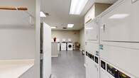 Located at 410 W 7th Street in downtown Fort Worth, Texas, this shared laundry facility offers a well-organized and functional space designed to accommodate multiple users efficiently. The single-floor layout is illuminated by a series of recessed skylight panels in the ceiling, providing bright, even lighting throughout the room. Neutral-toned walls and durable low-pile carpeting and concrete flooring contribute to a clean, utilitarian aesthetic.

The facility is equipped with a robust lineup of commercial-grade appliances, including multiple front-load and top-load washing machines, as well as stacked dryer units arranged along the perimeter walls to maximize usable floor space. A utility sink is installed along one wall, offering added convenience for pre-treatment or hand-washing needs. Ample folding counter space is provided along a built-in white ledge, making laundry sorting and folding straightforward.

A CSC payment kiosk is prominently positioned near the entrance, enabling cashless payment for machine use. A wooden bench at the center of the room offers a comfortable waiting area. Safety equipment, including a fire extinguisher, is visibly mounted on the wall. Decorative flower arrangements add a welcoming touch to an otherwise practical space. The facility also features a bulletin board and a security camera, reflecting attention to both communication and resident safety.