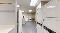Located at 410 W 7th Street in downtown Fort Worth, Texas, this shared laundry facility offers a well-organized and functional space designed to accommodate multiple users efficiently. The single-floor layout is illuminated by a series of recessed skylight panels in the ceiling, providing bright, even lighting throughout the room. Neutral-toned walls and durable low-pile carpeting and concrete flooring contribute to a clean, utilitarian aesthetic.

The facility is equipped with a robust lineup of commercial-grade appliances, including multiple front-load and top-load washing machines, as well as stacked dryer units arranged along the perimeter walls to maximize usable floor space. A utility sink is installed along one wall, offering added convenience for pre-treatment or hand-washing needs. Ample folding counter space is provided along a built-in white ledge, making laundry sorting and folding straightforward.

A CSC payment kiosk is prominently positioned near the entrance, enabling cashless payment for machine use. A wooden bench at the center of the room offers a comfortable waiting area. Safety equipment, including a fire extinguisher, is visibly mounted on the wall. Decorative flower arrangements add a welcoming touch to an otherwise practical space. The facility also features a bulletin board and a security camera, reflecting attention to both communication and resident safety.