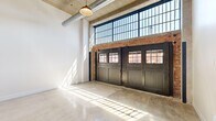 Located at 8 S Erie Street in Toledo's downtown district, this first-floor unit blends industrial character with modern finishes throughout its thoughtfully designed single-level layout. The most striking feature is the expansive main living area, where soaring ceilings, exposed concrete overhead, and visible silver ductwork establish a raw, industrial aesthetic. Oversized factory-style steel-framed windows run along the upper wall, flooding the space with natural light that reflects brilliantly off the polished concrete floors. A set of bold black carriage-style doors with windowed panels serves as a dramatic architectural focal point, framing street views while reinforcing the building's historic character. Exposed brick accents and original concrete columns further enhance the industrial ambiance.

The open kitchen features dark cabinetry, stainless steel appliances including a refrigerator, microwave, and range, and a central island with seating, all anchored by the same polished concrete flooring. Private rooms transition to soft carpeting underfoot, offering a quieter, more intimate atmosphere, with an exposed concrete column adding a rustic touch. The full bathroom is cleanly appointed with a dark-vanity cabinet, white countertop, industrial-style wall sconces, and a tub-shower combination, complemented by polished concrete flooring.