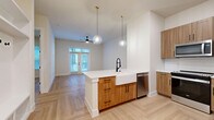 Located in Austin, TX (78756), this thoughtfully designed one-bedroom, one-bathroom residence offers a well-appointed layout with high-end finishes throughout. The open-concept kitchen and living area serves as the heart of the home, anchored by a striking center island topped with veined quartz countertops and a deep farmhouse sink with a matte black faucet. Warm-toned wood cabinetry with black hardware lines the kitchen walls, complemented by stainless steel appliances including a refrigerator and microwave. Globe pendant lights with brass fixtures hang above the island, adding a contemporary yet inviting touch. The living area flows seamlessly from the kitchen, featuring light herringbone-patterned hardwood flooring that extends throughout the entire residence. French doors flood the living space with natural light and open to a private balcony, while a ceiling fan adds comfort. The bedroom is a calm, well-lit retreat with large windows fitted with blinds and the same herringbone flooring. The full bathroom is finished with large-format white tile flooring, a wood vanity with quartz countertop, a framed mirror, matte black fixtures, and a soaking tub alongside marble-style tiled walls, creating a clean and modern aesthetic.
