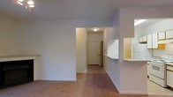 This single-floor unit in Fayetteville, North Carolina, offers a straightforward, functional layout designed for comfortable everyday living. The living area features neutral-toned carpet and smooth walls painted in a soft, light hue, creating a clean and understated atmosphere throughout. A small built-in shelf along one wall adds a subtle architectural detail and provides a convenient surface for display or storage.

A pass-through counter connects the living space to the kitchen, allowing for an open, communicative flow between the two areas. The kitchen, visible through the pass-through, includes white cabinetry and appliances, maintaining a bright and cohesive look. The counter-height pass-through also functions as a casual bar or serving space.

A central hallway leads toward the front entry door, which is fitted with a standard panel design and framed by warm overhead lighting. The hallway transitions between the main living space and the entryway, providing a clear and defined path through the unit. The flooring in the hallway shifts from carpet to a harder surface near the entry, offering a practical transition zone. Overall, the space presents a tidy, move-in-ready environment with a simple, no-frills design sensibility.
