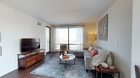 This single-floor residence combines modern finishes with functional design throughout its well-organized layout. The living room serves as the centerpiece of the home, featuring dark hardwood floors, large floor-to-ceiling windows dressed with sheer curtains, and direct access to a balcony offering sweeping city skyline views. Natural light fills the space generously, creating a bright and welcoming atmosphere.

The kitchen is thoughtfully designed with rich, dark wood cabinetry extending to the ceiling, granite-style dark countertops, and a full suite of stainless steel appliances including a refrigerator, range, and built-in microwave. A peninsula with bar seating connects the kitchen to the main living area, making it well-suited for casual dining and entertaining.

The bedrooms are comfortably sized, each featuring the same dark hardwood flooring, neutral walls, and ample natural light. One bedroom includes balcony access with city views, while another offers generous space for a desk and workspace area.

Two full bathrooms serve the residence. The primary bathroom includes a soaking tub, a rich wood vanity, and tile flooring, while the secondary bathroom features a tiled shower-tub combination and a clean, streamlined vanity. Both bathrooms maintain a consistent, neutral aesthetic throughout.