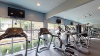 This fitness center is designed to support a comprehensive workout experience within a single-level layout that flows between two connected zones. The cardio area is lined with large windows that run nearly the full length of the wall, flooding the space with natural light and offering open views of the surrounding streetscape and greenery. Multiple treadmills, elliptical machines, and a stationary bike are arranged along this windowed wall, each equipped with individual display screens. Wall-mounted televisions provide additional entertainment options throughout the room. Full-length mirrors are installed along interior walls, visually expanding the space and allowing users to monitor their form. The strength training area occupies an adjoining section and includes a multi-station cable machine, a weight bench, a dumbbell rack with a range of free weights, and a stability ball. Rubber flooring throughout provides durability and cushioning. Recessed lighting illuminates the entire space evenly, supplementing the abundant natural light. A bulletin board and posted fitness center policies keep users informed. The light blue walls and clean, neutral finishes give the space a modern and welcoming atmosphere suited for a variety of fitness routines.