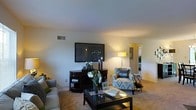 This single-floor space presents a thoughtfully arranged living area that balances functionality with a warm, homey aesthetic. The living room serves as the central gathering space, featuring wall-to-wall carpeting that adds a soft, comfortable feel underfoot. A large flat-screen television is mounted on the wall above a dark wood media console, creating a natural focal point for the room. Warm ambient lighting from a floor lamp casts a welcoming glow throughout the space, complementing the neutral wall tones. Seating includes a patterned accent chair accented with a decorative "HOME" pillow, alongside a sofa positioned to encourage conversation and relaxation. A sleek, dark glass-topped coffee table anchors the seating arrangement, topped with decorative books and accessories. Fresh white floral arrangements add a lively, natural touch to the room. A doorway visible in the background leads to an adjacent room, suggesting a connected and flowing floor plan. The space benefits from a clean, uncluttered layout with smooth ceilings and standard wall vents, reflecting a practical and comfortable living environment suitable for everyday use.