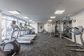 Located at 1810 Highpoint Ave in Richmond, Virginia, this fitness center is a comprehensively outfitted exercise facility designed to support a wide range of workout routines. The space is finished with durable patterned carpet flooring throughout and illuminated by recessed flush-panel ceiling lights, creating a bright and energetic environment. Bold red accent walls punctuate the interior, adding a dynamic visual element that complements the facility's active purpose.

The cardio zone is generously appointed with multiple treadmills, stair climbers, and elliptical machines, many equipped with individual display screens. Large windows along the perimeter walls allow natural light to fill the space and provide views of the surrounding neighborhood. A Peloton cycling bike and additional stationary bikes are available for low-impact cardio training.

The strength and functional training area features a Synrgy360 multi-functional cable and resistance system, a variety of free weights, medicine balls, and a dedicated barre rail mounted along the wall. A large flat-screen television is positioned for easy viewing during workouts. The facility also includes a separate, more intimate alcove with a treadmill and cycling bike alongside wide windows, offering a quieter training option. The overall layout is open and well-organized, accommodating multiple users simultaneously with ease.