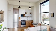 Located in Chicago's vibrant Uptown neighborhood, this single-floor unit at 4700–4766 N Clifton Ave offers a thoughtfully designed open-concept layout that seamlessly blends the kitchen, dining, and living areas. Polished concrete floors run throughout, lending an industrial-modern aesthetic that pairs well with the clean white walls and crisp black window frames.

The kitchen is well-appointed with stainless steel appliances—including a gas range and full-size refrigerator—white subway tile backsplash, a combination of white upper cabinetry and warm wood-toned lower cabinets, and open shelving for display. Pendant lighting anchors the space with a contemporary touch. A compact dining area with a round table sits adjacent, positioned to take advantage of the large windows.

The living area is bathed in natural light from oversized, black-framed windows that frame sweeping views of the Chicago skyline and Lake Michigan beyond, providing a striking visual backdrop. The bedroom is a calm, minimalist retreat with neutral tones and clean lines. The bathroom features a glass-enclosed walk-in shower with white tile surround and sleek matte black fixtures, adding a refined contrast. Overall, the unit balances modern industrial design with warmth and functionality.