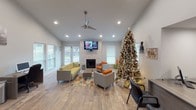 This well-appointed single-floor space offers an open, airy layout highlighted by vaulted ceilings and recessed lighting throughout. The central living area serves as the focal point, anchored by a striking floor-to-ceiling stacked stone fireplace with a wall-mounted flat-screen TV above it. Ample natural light pours in through multiple windows fitted with white blinds, illuminating the warm wood-look flooring that runs seamlessly across the space. Comfortable, modern seating in bold accent colors — including yellow and orange — is arranged around a colorful area rug, fostering an inviting atmosphere for gatherings. A ceiling fan adds both style and function overhead. Adjacent to the living area, a compact kitchenette features rich dark-wood cabinetry, white countertops, a stainless steel sink, and a beverage refrigerator — ideal for hosting. Soaring ceilings above the kitchenette amplify the sense of openness. A dedicated workspace with a desk and computer is tucked into a corner of the main room, offering a practical area for productivity. The layout flows naturally between zones, making the space equally suited for professional, social, or community use.