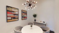 This thoughtfully designed single-floor space blends contemporary aesthetics with functional comfort. The interior immediately draws attention with a statement sputnik chandelier — a multi-arm pendant light with exposed Edison bulbs set against a dark bronze finish — that serves as a bold focal point overhead. The walls are finished in a clean, bright white, amplifying the sense of openness and allowing the colorful abstract artwork displayed in black frames to stand out vividly. The framed pieces feature dynamic horizontal bands of color, adding an energetic, artistic character to the space. A round white table at the center is flanked by soft grey upholstered chairs, creating an intimate and welcoming gathering area. A small potted succulent on the table adds a subtle natural touch. Along one wall, a sleek console or credenza supports a collection of small decor items and what appears to be informational materials, suggesting this space may serve a professional or hospitality function. A lush potted tropical plant in the corner further enlivens the room with greenery. The overall ambiance is clean, modern, and curated — striking a balance between approachable warmth and polished design sensibility.