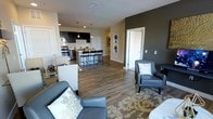 This single-floor residence showcases a thoughtfully designed open-concept layout that effortlessly connects the living area and kitchen into one cohesive, flowing space. Warm hardwood-style flooring runs throughout, lending a sense of continuity and warmth to the entire floor plan.

The living area is generously proportioned, featuring stylish gray upholstered seating arranged over a patterned area rug. A bold accent wall in deep olive-green adds a striking focal point and visual depth to the room. Recessed lighting overhead ensures the space is well-lit, complementing the overall modern aesthetic.

The kitchen is a standout feature, designed with dark espresso cabinetry contrasted against crisp white countertops and a clean white backsplash. Stainless steel appliances add a professional touch, while pendant lights hang above a central island with bar-height seating — an ideal setting for casual dining or entertaining. The kitchen's open sightlines to the living space make it both functional and social.

A hallway with two white paneled doors leads to additional private rooms, providing clear separation between the living areas and more secluded spaces. The overall ambiance is contemporary and refined, balancing neutral tones with bold accents for a polished, inviting atmosphere.