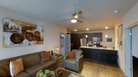 This single-floor space showcases an open-concept layout that seamlessly connects the living area and kitchen, creating a cohesive and functional environment. The living area is anchored by a ceiling fan with an integrated light fixture and features warm hardwood-style flooring that extends throughout the space. Comfortable seating, including a sofa and armchair, is arranged to encourage conversation, while decorative artwork and accent lighting add character to the room.

The kitchen is a standout feature, equipped with dark espresso cabinetry that extends to the upper walls, stainless steel appliances including a refrigerator, microwave, and range, and a light-toned countertop that contrasts elegantly with the darker elements. Pendant lights hang above the kitchen island, which also serves as a breakfast bar with bar seating, making it ideal for casual dining or entertaining.

A stacked washer and dryer unit is conveniently tucked into a dedicated closet space, adding practicality to the layout. Recessed lighting throughout ensures the space is well-lit, contributing to a warm and inviting ambiance. The overall design balances modern aesthetics with comfortable, everyday functionality.