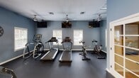 This dedicated fitness facility is thoughtfully designed to support a comprehensive workout experience within a single, open floor plan. The space is finished with cool blue-gray walls and durable dark rubber flooring, creating a clean, energizing atmosphere conducive to exercise.

The cardio zone is generously equipped with treadmills, an elliptical trainer, and cycling bikes, each positioned facing individual wall-mounted flat-screen televisions for an engaging workout. Ceiling fans overhead ensure comfortable air circulation throughout this area. Natural light filters in through multiple white-framed windows fitted with horizontal blinds, offering a bright and inviting environment.

The strength training section features a multi-function cable machine alongside a well-stocked rack of exercise balls, resistance bands, foam rollers, and medicine balls in a variety of sizes. Full-length mirrored walls in this zone expand the sense of space and allow users to monitor their form during workouts. Overhead fluorescent lighting and an additional ceiling fan keep this area well-lit and ventilated.

A pass-through window connects to an adjacent room, and a French door with glass panes provides convenient access between areas. A wall-mounted hand sanitizer dispenser and a step-on waste bin reflect thoughtful attention to cleanliness and functionality. Overall, the space is practical, well-appointed, and designed to accommodate a wide range of fitness activities.