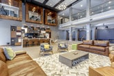 This contemporary communal space is thoughtfully designed to serve as both a welcoming lobby and a social hub. The layout is open and generously proportioned, with warm brick-pattern tile flooring running throughout, anchoring the space with a grounded, urban feel.

A standout feature is the built-in coffee bar, equipped with a professional espresso machine and outfitted with a modular wood-and-metal shelving unit displaying decorative accents and coffee service essentials. Bar stools line the counter, creating an inviting spot for casual interaction. Dramatic dome pendant lights in dark tones with copper interiors hang overhead, adding a refined, modern touch.

The lounge area is furnished with a mix of leather sofas and contemporary chairs in earthy and cool-toned hues, centered around a low coffee table atop a patterned area rug. A large-scale blue wall mural adds visual depth and character to the seating nook.

Soaring ceilings with exposed wood accents and an upper-level glass-railed mezzanine give the space a dramatic sense of vertical scale. Floor-to-ceiling windows flood the interior with abundant natural light and provide direct views of an outdoor courtyard and surrounding streetscape. Glass doors provide seamless indoor-outdoor connectivity.