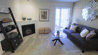 This single-floor space centers around a well-appointed living area that balances warmth and functionality. A built-in electric fireplace serves as a focal point, framed by a clean white mantel that provides display space and adds a classic architectural touch. The room is finished with neutral wall-to-wall carpeting, contributing to a soft, inviting atmosphere throughout.

Natural light is a defining feature of the living area, delivered through a large sliding glass door that opens onto a private balcony or patio. The door is dressed with sheer white curtains, allowing light to filter in gently while maintaining a bright and airy feel. The outdoor view of mature trees adds a natural backdrop that enhances the sense of tranquility.

The layout accommodates comfortable seating arrangements, with ample room for a sofa and accent pieces, centered around a round glass-top coffee table that adds an open, uncluttered quality to the space. Neutral tones throughout the walls and flooring create a versatile canvas adaptable to a variety of furnishing styles. Overall, the space offers a practical yet welcoming environment suited for relaxed living.