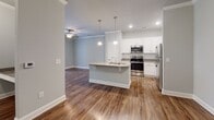 Located along US-64 E in Pittsboro, NC, this single-story unit offers a well-designed, open-concept layout that seamlessly connects the kitchen, dining, and living areas. The kitchen is a standout feature, showcasing white shaker-style cabinetry, granite countertops, a subway tile backsplash, and a full suite of stainless steel appliances including a range, microwave, refrigerator, and dishwasher. A central island with a granite surface and undermount sink provides additional workspace and casual seating, illuminated by two pendant lights hanging overhead. Recessed lighting and crown molding add a polished finish to the main living area, which is further complemented by rich hardwood-style flooring and a ceiling fan. The bedrooms are carpeted for comfort and feature ceiling fans, ample natural light from sizable windows with blinds, and neutral tones throughout. The bathroom is appointed with dark-stained cabinetry, a granite vanity top, a tub-shower combination, and tile flooring. Soft gray walls flow consistently through the entire unit, creating a cohesive, modern aesthetic.