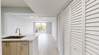 This well-appointed one-bedroom, one-bathroom unit offers a clean, modern aesthetic throughout its efficiently designed single-floor layout. Light-toned hardwood flooring runs continuously across the living spaces, complementing the soft gray walls and crisp white trim that define the interior palette.

The combined kitchen and living area forms the heart of the unit. The kitchen is outfitted with light wood cabinetry extending to the ceiling, stone countertops, a deep stainless steel sink, and a full suite of stainless steel appliances including a refrigerator, range, and over-the-range microwave. A peninsula countertop provides a natural division between the kitchen and the open living area, which benefits from sliding glass doors leading to a private balcony overlooking palm-lined surroundings and the urban streetscape beyond.

The bedroom is a generously proportioned retreat featuring large windows that invite abundant natural light and offer outdoor views. Exposed ceiling beams add a subtle architectural detail to the space. A dedicated closet provides practical storage.

The bathroom is finished in a clean, contemporary style with white subway tile surrounding a full bathtub and shower combination, light flooring, and simple chrome fixtures. A hallway connects the main living areas, completing the functional and cohesive floor plan.