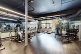 Located at 1050 Blanche St in West Palm Beach, FL, this fitness center offers a thoughtfully designed workout space that balances functionality with a modern industrial aesthetic. The single open-plan floor features rich wood-look flooring throughout, providing a warm contrast to the dark, exposed ceiling that showcases structural beams, ductwork, and linear LED lighting — all hallmarks of a contemporary industrial design.

The cardio zone is well-appointed with multiple treadmills, elliptical trainers, and a stationary bike, positioned to take advantage of natural light streaming in through a row of large windows that frame outdoor greenery. Floor-to-ceiling mirrors line key sections of the space, visually expanding the room and allowing users to monitor their form during workouts.

The strength training area is equally comprehensive, featuring a multi-functional cable machine, a full dumbbell rack, weight benches, and additional resistance equipment. A dedicated functional fitness corner houses medicine balls and stability balls on organized storage racks, catering to a variety of training styles. The space concludes with a glass-door entry area and access to additional rooms, ensuring smooth circulation throughout. Overall, this fitness center delivers a clean, energizing environment suited for a complete range of workout routines.