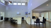 This single-floor space makes an immediate impression with its dramatic vaulted ceilings, supported by exposed white beams that draw the eye upward. A generous row of clerestory windows runs along the upper wall, flooding the interior with abundant natural light and creating an airy, open atmosphere throughout. The walls are finished in a soft neutral gray, contributing to the clean, contemporary aesthetic.

At the heart of the space is a well-designed wet bar and counter area featuring light granite countertops atop white cabinetry, a built-in beverage refrigerator, and a bar-height counter lined with sleek black bar stools. A wall-mounted flat-screen display and decorative shelving with framed artwork add functional and visual interest to this entertaining zone. The flooring throughout is a rich, dark wood-tone tile that grounds the bright, open interior with warmth and contrast.

An adjacent area, visible through a wide arched passageway, reveals a workspace or reception area equipped with a desktop computer and additional counter space, with bright, naturally lit rooms extending further beyond. A classic round-back accent chair anchors a transitional corner near the passageway. Together, these elements create a cohesive, inviting space that balances function with refined design.