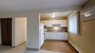 This single-floor residence offers a practical and well-organized layout designed for comfortable everyday living. The kitchen is a standout feature, outfitted with an abundance of white raised-panel cabinetry that provides generous storage from floor to ceiling. A double-basin sink sits beneath the upper cabinets, accompanied by a full suite of white appliances including a refrigerator, electric range, and under-cabinet range hood. Dark-toned countertops offer a subtle contrast against the bright white cabinetry, while warm hardwood-style flooring grounds the kitchen space with a clean, polished look. Natural light filters into the kitchen area through a large window dressed with vertical blinds, brightening the room with a soft, diffused glow. A baseboard heating unit runs along the wall, providing efficient climate control. The adjoining areas transition to wall-to-wall carpeting, offering a softer, more relaxed feel underfoot. Neutral warm tones on the walls lend a cohesive and inviting ambiance throughout. The open sightline between the kitchen and adjacent rooms creates a sense of connectivity and ease of movement, making the overall layout both intuitive and livable.