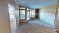 This single-floor residence offers a thoughtfully designed layout that balances openness with defined living spaces. The expansive living room is a standout feature, showcasing elegant crown molding, soft wall-to-wall carpeting, and a generous arrangement of windows with transom lights above that flood the space with natural light. Built-in shelving units flank the exterior wall, providing both display and storage opportunities, while a door leads to an outdoor area. A unique architectural niche in the wall adds a decorative touch and connects the living area to the rest of the unit. The kitchen is well-equipped with rich dark wood cabinetry, granite countertops, stainless steel appliances including an electric range and over-the-range microwave, and a peninsula that opens to the main living space via track lighting overhead. The bedroom is a comfortable retreat, featuring plush carpeting, a ceiling fan with light fixture, and a wide window that brings in natural light and outdoor views. The bathroom is finished with warm wood-toned cabinetry, a granite-topped vanity, a large mirror, and tiled shower. A spacious walk-in closet with built-in shelving and hanging rods is accessible directly from the bathroom, offering ample storage. Throughout, crown molding and neutral tones create a cohesive, inviting ambiance.