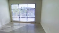 This single-level space presents a clean, modern aesthetic defined by its straightforward yet functional design. The most prominent feature is the large, multi-pane floor-to-ceiling window that spans a significant portion of one wall, fitted with horizontal blinds and offering views of mature trees and neighboring structures. This generous glazing floods the interior with natural light, casting bright reflections across the polished concrete flooring below. The concrete floors contribute to the space's contemporary, minimalist character while providing a durable and low-maintenance surface. The walls are finished in a smooth, neutral tone that complements the industrial quality of the flooring. Clean white baseboards provide a subtle contrast and define the room's perimeter. The overall ambiance is open, uncluttered, and versatile, making it well-suited for a variety of uses. The combination of abundant natural light, polished floors, and neutral finishes creates a bright and practical environment with a modern edge.