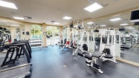 Located at 6001 SW 70th St in South Miami, FL, this fitness center offers a well-appointed, single-floor workout facility designed to support a comprehensive exercise routine. The space is thoughtfully laid out across its compact footprint, maximizing functionality without sacrificing comfort or openness.

The room features a durable rubber flooring surface ideal for high-traffic fitness use, and the warm beige walls are accented by large, arched windows along one side that flood the space with natural light and provide views of lush tropical greenery and an adjacent pool area. Ceiling-mounted recessed panel lighting ensures bright, even illumination throughout.

The equipment selection is extensive for the space's size. Cardiovascular machines include multiple treadmills and a stationary recumbent bike, while a dedicated row of free weights and a dumbbell rack caters to strength training needs. Several multi-use cable and resistance machines — including HOIST and Cybex branded units — line the room, enabling a wide range of strength and conditioning exercises. Wall-mounted mirrors on multiple sides visually expand the space and assist with form during workouts. Ceiling-mounted televisions are positioned for easy viewing from various equipment stations.

Entry is provided through elegant glass-panel double doors, and posted fitness rules signage reflects a well-managed, community-oriented facility. Overall, the space delivers a clean, bright, and fully functional fitness environment.