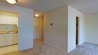 This single-floor residential space offers a clean, functional layout designed for comfortable everyday living. The interior is finished with wall-to-wall carpeting in a neutral tone, contributing to a warm and quiet atmosphere throughout. The walls are painted in a soft, light cream hue, creating a bright and open feel that makes the space feel welcoming and easy to personalize.

A defined entryway leads into the main living area, separated by a partial wall that adds a sense of structure and delineation between spaces. The entry corridor is equipped with a ceiling-mounted flush light fixture, providing practical illumination upon arrival. A standard entry door with simple hardware serves as the main point of access, reinforcing the straightforward, no-frills character of the space.

Electrical outlets and light switch panels are positioned at standard heights along the walls, ensuring convenience throughout. The layout benefits from an open flow between the entry and the adjoining rooms, making navigation intuitive and efficient. The overall design is modest and utilitarian, prioritizing function while maintaining a tidy, neutral aesthetic that suits a wide range of furnishing styles. The single-floor configuration ensures easy accessibility and a cohesive living experience without the need to navigate between levels.