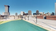 Located atop a building in the heart of New Orleans, this rooftop dog run offers a well-appointed outdoor space designed specifically for pets and their owners. The open-air area is surfaced with vibrant green artificial turf, providing a clean and comfortable environment for dogs to move freely. A retractable or fixed canopy structure extends over a slatted deck platform, offering shaded relief from the Louisiana sun while maintaining an open, airy feel. The space is fully enclosed by secure metal fencing and features a gated entry, ensuring a safe environment for off-leash activity. Waste disposal stations are conveniently positioned throughout. Access is provided through a key-controlled entry door, adding an additional layer of security. The rooftop setting delivers breathtaking, unobstructed panoramic views of the New Orleans Central Business District skyline, with prominent downtown high-rises visible in every direction under wide-open blue skies. The elevated vantage point creates a striking contrast between the urban landscape and the pet-friendly amenity space. This thoughtfully designed rooftop dog run combines practical functionality with an exceptional urban outdoor experience for both pets and residents.