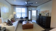 This single-floor space centers around a well-appointed living area that balances comfort and functionality. Large windows dressed with horizontal blinds and patterned curtains allow abundant natural light to fill the room, creating a bright and welcoming atmosphere. A ceiling fan mounted overhead provides both airflow and a subtle architectural focal point.

The living area is anchored by a plush white sofa adorned with decorative pillows, complemented by a pair of dark accent chairs arranged near the windows. A substantial leather ottoman serves as a central coffee table, resting atop a woven area rug that defines the seating zone against dark hardwood flooring. A tray with decorative items sits atop the ottoman, adding a curated touch.

A dark, multi-drawer dresser positioned along the right wall provides generous storage and is topped with a potted plant, adding a natural element to the space. Recessed lighting supplements the natural light throughout. An open doorway leads to an adjacent room, suggesting a connected and flowing floor plan. Overall, the space presents a cohesive, casually refined aesthetic with warm tones and layered textures.