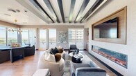 Located at 2351 Plaza Blvd in Richardson, Texas, this single-floor sky lounge is an elegantly designed space that seamlessly integrates a living room, dining room, and kitchen into one cohesive, open layout. The living area serves as the centerpiece of the space, anchored by a striking linear electric fireplace set beneath a framed wall-mounted television, flanked by warm-toned built-in shelving with integrated lighting. Bold dark ceiling beams add architectural depth and contrast against the light, neutral walls, while wide-plank hardwood flooring runs throughout, lending warmth and continuity. Floor-to-ceiling glass doors and expansive windows flood the interior with abundant natural light and provide direct access to an outdoor balcony, where panoramic views of the Richardson skyline and surrounding landscape can be enjoyed. The dining area is distinguished by a modern round table beneath a sculptural gold chandelier, positioned to take full advantage of the sweeping corner views through glass balcony doors. The kitchen features a generous white quartz waterfall island with bar seating, wood cabinetry, and a trio of gold pendant lights, completing the refined and cohesive aesthetic of this elevated lounge space.