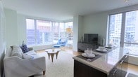 This single-floor residence offers an open-concept layout that combines the kitchen, dining, and living areas into one cohesive, light-filled space. Floor-to-ceiling windows wrap around the living area, flooding the interior with natural light and framing dynamic city skyline views, creating a strong connection between the interior and the urban environment outside.

The living area features light hardwood flooring, neutral walls, and ample room for comfortable seating arrangements. A large area rug anchors the space, complementing the modern furnishings and adding warmth to the bright, airy atmosphere.

The kitchen is positioned at the edge of the open-plan area, defined by a granite-topped peninsula that serves as both a workspace and a casual dining counter. Dark cabinetry contrasts elegantly with the light stone countertops, lending a contemporary, refined aesthetic.

Overall, the space exudes a clean, modern ambiance characterized by an abundance of natural light, high-rise urban views, and a thoughtful open layout that maximizes both functionality and visual appeal.