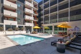 Located at 417 Centinela Ave in Inglewood, California, this outdoor amenity space serves as the social centerpiece of a modern multi-story residential community. The courtyard is designed to maximize the Southern California lifestyle, anchored by a generously sized swimming pool with crystal-clear water and integrated in-pool lighting. Adjacent to the pool, a dedicated spa provides a relaxing retreat. The surrounding deck is finished in clean concrete, thoughtfully furnished with rows of sleek lounge chairs, side tables, and bold yellow market umbrellas that add a vibrant pop of color against the contemporary architectural backdrop. A separate lounge seating area offers a comfortable space for socializing, complete with patterned accent rugs and colorful throw pillows. Lush landscaping, including agave plants and layered greenery, lines the raised planters and softens the perimeter of the space. The courtyard is enclosed on multiple sides by the residential building's facade, which features a striking interplay of gray concrete panels, white balconies, and bold orange and red accent walls, creating an energetic and contemporary visual identity. The open-air design allows abundant natural light to flood the space throughout the day, making this courtyard an inviting and dynamic outdoor destination.