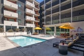Located at 417 Centinela Ave in Inglewood, California, this outdoor amenity space serves as the social centerpiece of a modern multi-story residential community. The courtyard is designed to maximize the Southern California lifestyle, anchored by a generously sized swimming pool with crystal-clear water and integrated in-pool lighting. Adjacent to the pool, a dedicated spa provides a relaxing retreat. The surrounding deck is finished in clean concrete, thoughtfully furnished with rows of sleek lounge chairs, side tables, and bold yellow market umbrellas that add a vibrant pop of color against the contemporary architectural backdrop. A separate lounge seating area offers a comfortable space for socializing, complete with patterned accent rugs and colorful throw pillows. Lush landscaping, including agave plants and layered greenery, lines the raised planters and softens the perimeter of the space. The courtyard is enclosed on multiple sides by the residential building's facade, which features a striking interplay of gray concrete panels, white balconies, and bold orange and red accent walls, creating an energetic and contemporary visual identity. The open-air design allows abundant natural light to flood the space throughout the day, making this courtyard an inviting and dynamic outdoor destination.
