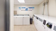 Located at 2417 Hurontario St in Mississauga, Ontario, this shared laundry facility is a well-organized, functional space designed to serve building residents efficiently. The room features a drop ceiling with recessed lighting panels that provide bright, even illumination throughout. The flooring consists of neutral beige ceramic tiles, which extend partway up the walls in a matching tile surround, lending the space a clean and easy-to-maintain finish. The remaining walls are finished in a light, neutral tone that keeps the area feeling open and bright. The facility is equipped with multiple top-loading commercial washers and front-loading dryers from Huebsch, all coin-operated and clearly numbered for easy identification. A utility sink is also present, offering added convenience for pre-treating garments. Instructional signage and operating guides are posted prominently on the walls, ensuring straightforward use of all machines. A dedicated notice board near the entrance provides residents with relevant building information. The room is accessed through a solid door and includes a waste bin for general tidiness. Overall, this laundry room is a practical, well-maintained shared amenity that prioritizes functionality and ease of use for building residents.
