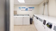 Located at 2417 Hurontario St in Mississauga, Ontario, this shared laundry facility is a well-organized, functional space designed to serve building residents efficiently. The room features a drop ceiling with recessed lighting panels that provide bright, even illumination throughout. The flooring consists of neutral beige ceramic tiles, which extend partway up the walls in a matching tile surround, lending the space a clean and easy-to-maintain finish. The remaining walls are finished in a light, neutral tone that keeps the area feeling open and bright. The facility is equipped with multiple top-loading commercial washers and front-loading dryers from Huebsch, all coin-operated and clearly numbered for easy identification. A utility sink is also present, offering added convenience for pre-treating garments. Instructional signage and operating guides are posted prominently on the walls, ensuring straightforward use of all machines. A dedicated notice board near the entrance provides residents with relevant building information. The room is accessed through a solid door and includes a waste bin for general tidiness. Overall, this laundry room is a practical, well-maintained shared amenity that prioritizes functionality and ease of use for building residents.