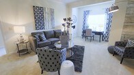 This single-floor space showcases a thoughtfully designed interior with a refined, contemporary aesthetic. The living area is anchored by a plush gray sofa accented with patterned throw pillows, complemented by a bold geometric accent chair and a sleek mirrored coffee table that adds a touch of glamour. A dark, textured area rug grounds the seating arrangement, while a decorative white garden stool provides a versatile accent piece. Large-scale wall art in silver, white, and charcoal tones adds visual depth and personality to the neutral walls. The space flows seamlessly into a secondary area, likely used as a dining or workspace, featuring a desk and chairs set beneath a large window dressed with bold black-and-white patterned curtains that frame an abundance of natural light. The light-colored carpeted flooring runs throughout, contributing to the cohesive and airy ambiance of the space. Decorative floral arrangements and metallic accents are thoughtfully placed throughout, reinforcing the sophisticated, monochromatic style. Overall, the space offers a well-balanced blend of comfort and contemporary design sensibility.