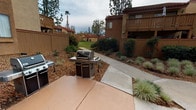 This residential community exterior showcases a thoughtfully designed, single-story ground-level environment set within a multi-building complex. The architecture features warm beige stucco facades, red accent doors, and classic red-tiled rooflines, lending a cohesive Mediterranean-influenced aesthetic throughout the property.

Gated, fenced walkways lined with ground cover plantings and ornamental shrubs connect the various buildings, creating a sense of privacy and separation between units. Mature pine and palm trees are distributed across the grounds, providing natural shade and an established, leafy atmosphere. Lush lawn areas open up between buildings, offering green communal space framed by well-maintained landscaping beds with mixed foliage and bark mulch borders.

An outdoor patio area is equipped with full-size gas grills, making it well-suited for communal gatherings and outdoor cooking. Concrete pathways wind through the property, connecting building entrances and common spaces. Black metal stair railings with granite-finish treads provide access between ground-level and upper-floor units, complementing the building's stucco exteriors.

Ground-floor unit entryways are flanked by low-growing ground cover and accented with wall-mounted lantern-style fixtures. Upper-floor balconies with metal railings overlook the landscaped interior courtyard, completing a community that balances functional outdoor living with attractive, park-like surroundings.