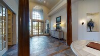This sophisticated single-floor space makes a striking first impression with its grand foyer, where soaring ceilings and oversized arched French doors flood the entry with natural light. The entryway is anchored by distinctive stone tile flooring in a bold geometric pattern, accented with warm-toned inlays, and flanked by rich dark wood columns that add architectural gravitas. A refined console table with framed artwork and wall sconces completes the welcoming aesthetic.

Beyond the foyer, the main lounge area exudes classic elegance with soaring ceilings, a dramatic wrought-iron chandelier, and a limestone-accented fireplace surround serving as the room's focal point. Plush blue velvet sofas, patterned armchairs, and a central storage-style coffee table are arranged over a deep blue area rug, creating a warm and inviting gathering space. Tall arched mirrors flank the fireplace, amplifying the sense of grandeur.

A well-appointed kitchenette off the main area features granite countertops, white cabinetry with open display shelving, subway tile backsplash, a built-in beverage refrigerator, and a coffee station — ideal for entertaining or everyday hospitality.

A carpeted office or private sitting room offers a quieter retreat, furnished with a tufted wingback chair, a glass coffee table, and a full-length mirror, with direct access to an outdoor area through a glass-paned door.