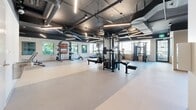 Located at 3488 Ketcheson Rd in Richmond, British Columbia, this well-appointed fitness facility offers a generously sized, single-floor exercise room designed to support a full range of workout activities. The space is defined by its industrial-modern aesthetic, featuring an exposed dark ceiling with visible ductwork and HVAC systems, complemented by sleek linear LED lighting that keeps the environment bright and energizing.

Large windows line multiple walls, flooding the interior with natural light and providing views of the surrounding neighbourhood. The durable, light-toned flooring spans the entire area and is well-suited for both equipment use and open floor exercises.

The facility is thoughtfully zoned into distinct workout areas. A dedicated strength training section houses cable machines, a multi-functional rack system, weight benches, and free weight storage. The cardio zone features treadmills, elliptical machines, a stationary bike, and stability ball storage. A more open, flexible area provides ample room for stretching or group activity.

A vibrant geometric mural in blue, green, orange, and red adds a dynamic visual accent, while practical amenities including a water fountain station and restroom facilities ensure user comfort. Wall-mounted monitors enhance the workout experience throughout the space.