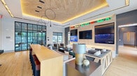 This contemporary single-floor amenity facility offers a thoughtfully designed environment across four distinct areas. The entrance hallway sets an inviting tone with soft carpeting, warm wood-paneled ceilings with recessed amber lighting, built-in shelving, and clean white walls accented by textured tile details. A natural wood bench and large glass doors allow abundant natural light to fill the transitional space. The expansive living lounge is a standout feature, boasting a striking natural wood-plank ceiling with warm cove lighting and bold circular pendant fixtures. Multiple seating arrangements with plush gray sofas surround a large wood-paneled media wall lined with multiple flat-screen televisions and a colorful LED ticker display. A built-in bar-height counter with leather stools adds a social dimension, while floor-to-ceiling black-framed glass walls and windows connect the interior to outdoor views. Adjacent to the lounge, a café-style kitchen and dining area features a herringbone-patterned hardwood floor, a sleek white waterfall island with a sink, dramatic teardrop pendant lighting, and an array of round dining tables with modern chairs. Bold artwork and a dark feature wall anchor the space with character. The fitness center impresses with soaring ceilings, expansive floor-to-ceiling windows overlooking the surrounding area, vibrant green accent walls, and a full suite of cardio equipment on durable rubber flooring.