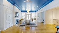 Eagle Rock at Freehold is a thoughtfully designed single-floor community space that blends bold aesthetics with a warm, welcoming atmosphere. The main gathering room immediately captures attention with its striking cobalt blue ceiling, which creates a dramatic contrast against the crisp white walls and wainscoting. Gleaming hardwood floors run throughout, adding warmth and continuity to the open layout. A central dining area anchors the room, surrounded by a mix of casual seating including playful hanging egg chairs that lend a relaxed, resort-inspired feel. A classic white fireplace with a decorative mantle serves as a charming focal point along one wall, complemented by a wall-mounted television for entertainment. Natural light pours in through sets of French doors, creating a bright and airy environment. Overhead, a cluster pendant light fixture and a ceiling fan add both style and function to the space. The open flow between areas makes the layout ideal for social gatherings and community events, while the eclectic mix of furnishings and layered area rugs contribute to an inviting, lively ambiance throughout.