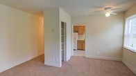 This single-floor residence offers a straightforward and functional layout connecting the main living area to the kitchen. The living space is finished with neutral wall-to-wall carpeting and light-toned walls, creating a clean and understated atmosphere. A ceiling fan is positioned overhead, contributing to the room's practicality and comfort.

A built-in shelving unit with a bifold door is integrated into a partial wall divider, providing convenient storage directly within the living area. This architectural element also helps define the transition between the living space and the kitchen beyond.

The kitchen is accessed through a wide doorway and features warm honey-toned wood cabinetry along both the upper and lower sections of the walls. A white refrigerator and countertop workspace are visible, and the flooring transitions to a light-colored tile with a subtle pattern, offering a durable and easy-to-clean surface. The kitchen benefits from its own overhead lighting, keeping the space bright and functional.

Overall, the residence presents a modest, well-organized layout with a neutral palette throughout, making it a practical and move-in ready space.