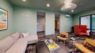 Located at 614 Rigsbee Ave in Durham, NC, this single-floor space offers a thoughtfully designed interior that blends contemporary style with functional comfort. The main lounge area is defined by sage green walls and a matching painted ceiling, creating a cohesive, calming atmosphere throughout. A striking woven pendant light fixture serves as a focal point overhead, complementing the recessed lighting that ensures the space is well-lit at all times.

Large, black-framed floor-to-ceiling windows and additional standard windows flood the interior with natural light while offering views of the surrounding Durham cityscape and neighboring architecture. The patterned, multi-tone carpet adds visual texture and grounds the eclectic furnishings, which include a neutral sectional sofa, bold red high-back accent chairs, and warm amber lounge chairs arranged to encourage both conversation and relaxation.

Colorful abstract artwork adorns the walls, reinforcing the space's modern aesthetic. A glass-partitioned area visible from the main lounge suggests a dedicated workspace or meeting zone with its own seating. Additional adjoining rooms, accessible through open doorways, extend the usable area of the space. A wall-mounted HVAC unit provides climate control. The overall ambiance is vibrant, welcoming, and well-suited for collaborative or social use.