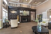 Located at 6115 Abbotts Bridge Road in Johns Creek, Georgia, this single-story community space presents a sophisticated and welcoming atmosphere defined by its refined architectural details and thoughtful design. The main lounge area commands attention with dramatically soaring coffered ceilings accented by recessed lighting, creating a sense of grandeur and openness. A striking dark-finished built-in fireplace surround serves as the room's focal point, flanked by display shelving and framed by bold architectural columns that add a classic, formal character. An abundance of natural light pours in through floor-to-ceiling French doors and transom windows, connecting the interior to the surrounding outdoor environment. The lounge is furnished with plush seating arrangements, including tufted sofas and wingback chairs, all set atop patterned carpeting that adds warmth and texture to the space. Wainscoting and chair rail molding run throughout, reinforcing the traditional aesthetic. A separate dining area features a sleek dark-finished table with upholstered seating and is adorned with decorative artwork and glass accent pieces. An elegant arched entryway leads to a foyer with a decorative tile floor, providing a gracious arrival experience. Together, these spaces offer a polished, comfortable environment ideal for gathering and socializing.