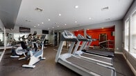 This fitness center is designed to support a comprehensive workout experience within a single-level layout. The space is finished with a durable rubber-speckled floor that provides traction and cushioning throughout the exercise areas. Recessed lighting illuminates the room evenly, complementing natural light that enters through several windows along one wall, offering an open and energizing atmosphere.

The cardio zone is prominently featured, housing multiple treadmills and stationary bikes arranged in organized rows. Elliptical and cross-trainer machines occupy a dedicated section, providing a variety of low-impact workout options. A strength training area includes a cable machine, weight bench, and a dumbbell rack stocked with a range of free weights, accommodating users at different fitness levels.

A bold orange accent wall serves as a focal point and adds visual energy to the space. Full-length mirrors line multiple walls, enhancing the sense of spaciousness while allowing users to monitor their form. A wall-mounted flat-screen television is positioned for visibility across the cardio area. The overall design balances functionality with a modern aesthetic, using a gray and orange color palette to create a motivating environment suited for daily fitness use.