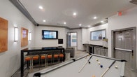 Located in Arlington, Texas, this contemporary single-floor amenity space is thoughtfully designed for entertainment and social gatherings. The open layout centers around a full-size billiards table, making it an ideal hub for recreation and leisure. A sleek bar counter with stylish orange upholstered stools provides a dedicated social gathering spot, complemented by a wall-mounted flat-screen television ideal for catching up on news or sports. Adjacent to the bar area, a comfortable lounge seating arrangement offers a relaxed space for conversation. The kitchenette along the far wall is well-appointed with gray cabinetry, dark countertops, a sink, and a built-in beverage refrigerator, ensuring guests have everything they need for entertaining. Large framed mirrors above the kitchenette counter enhance the sense of openness within the space. Throughout, recessed lighting in the smooth white ceiling provides bright, even illumination, while light wood-style flooring ties the space together with a clean, modern aesthetic. The neutral gray and white color palette, accented with chrome and contemporary finishes, creates a polished and inviting atmosphere suited for a variety of social activities.