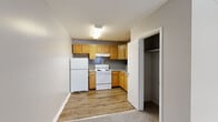 Located at 8223 Roosevelt Blvd in Philadelphia, PA, this single-floor unit offers an efficient and functional layout. The combined kitchen and living area makes smart use of the available space, with the kitchen featuring warm honey-toned wood cabinetry extending from floor to ceiling, providing ample storage. Grey speckled countertops line the L-shaped kitchen configuration, complemented by a stainless steel sink, a white electric coil range with an overhead exhaust hood, and a full-size refrigerator. The kitchen floor is finished with wood-look luxury vinyl plank, adding a clean, modern touch. Adjacent to the kitchen is a dedicated closet with a hanging rod, offering convenient storage. The living area is carpeted in a neutral grey tone and is bathed in natural light from a window fitted with white horizontal blinds. A wall-mounted air conditioning unit and baseboard heating ensure year-round comfort. The bathroom is clean and straightforward, featuring a full-size soaking tub with a tiled surround, a toilet, and a vanity. The overall color palette of soft greys and warm wood tones creates a cohesive, understated ambiance throughout the unit.