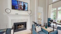 The Crowne at James Landing clubhouse presents a sophisticated, single-level gathering space defined by dramatic double-height ceilings and refined architectural detailing throughout. Tall, fluted columns frame the open lounge area, lending a stately character reinforced by wainscoting and paneled wall moldings finished in crisp white against warm neutral walls.

The main lounge serves as the centerpiece of the clubhouse, anchored by a classic white-mantled fireplace clad in stacked stone tile with a wall-mounted flat-screen television above. Comfortable teal upholstered armchairs with nailhead trim are arranged around low wood side tables, complemented by a generous taupe sofa and patterned area rug, creating an inviting seating arrangement suitable for both relaxing and socializing.

Floor-to-ceiling windows and glass-paned French doors flood the interior with abundant natural light while offering views of the surrounding greenery. Elegant two-toned drapery in cream and teal frames the large windows, adding a polished finishing touch. Wood-look plank flooring runs throughout, grounding the space with warmth and texture. Additional functional areas, accessible through interior French doors, provide private workspace and leasing office facilities, rounding out this well-appointed community hub.