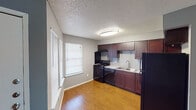 Located at 1647 Rutland Dr in Austin, Texas, this single-story unit offers a practical and well-appointed layout. The kitchen is a standout feature, showcasing rich dark-mahogany cabinetry that extends to the upper walls, providing generous storage. A gray subway tile backsplash complements the granite-style countertops, while black appliances — including a gas range, dishwasher, and refrigerator — complete the space. A double-basin stainless steel sink sits beneath a window that allows natural light to filter in. Warm wood-style flooring runs throughout the main living areas, adding a cohesive and inviting feel. The living space is open and functional, with neutral gray walls and a textured ceiling overhead. A ceiling fan is visible in one area, contributing to comfort year-round. The bathroom features a granite-topped vanity with a framed mirror, a toilet, and a tub/shower combination with a frosted privacy window. The hallway connects the living and bedroom areas efficiently, with the bedroom offering a carpeted floor and direct access to the bathroom. Blinds on the windows throughout the unit offer both privacy and light control, making this a practical and comfortable Austin home.